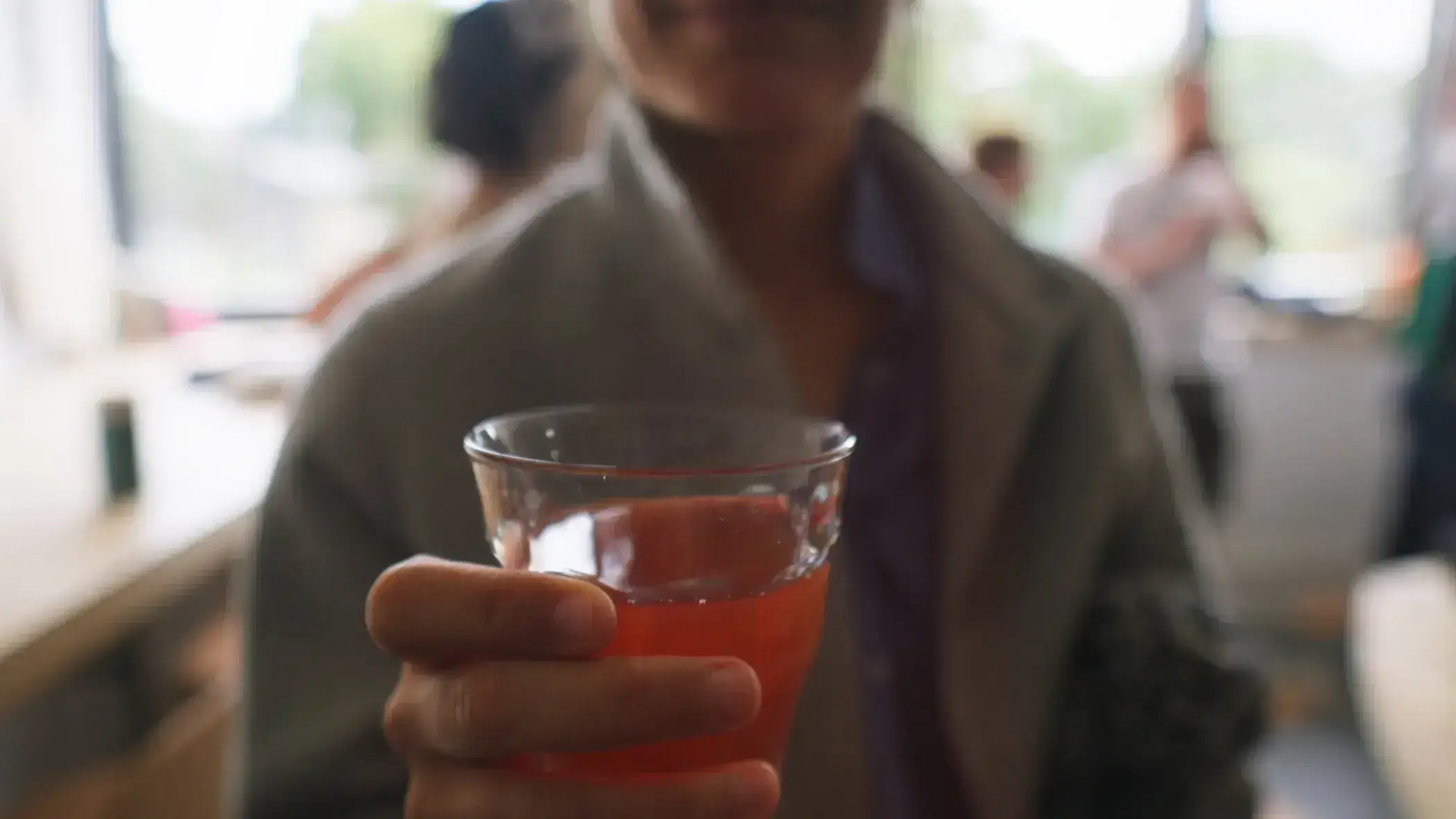 Person holding a glass of red drink, possibly Sindup, at an event with blurred people in the background.