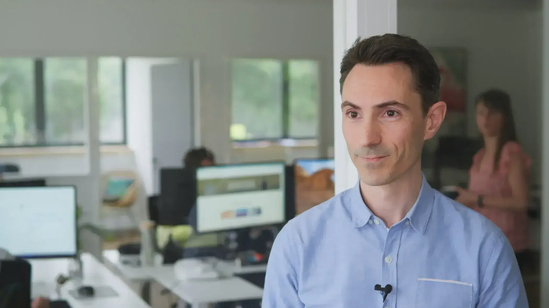 Man in a blue shirt in a modern office setting. Coworkers and computer screens visible in the background.