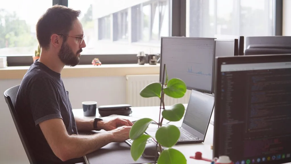 Man coding on a multi-monitor setup, possibly for Sindup, in a bright office.