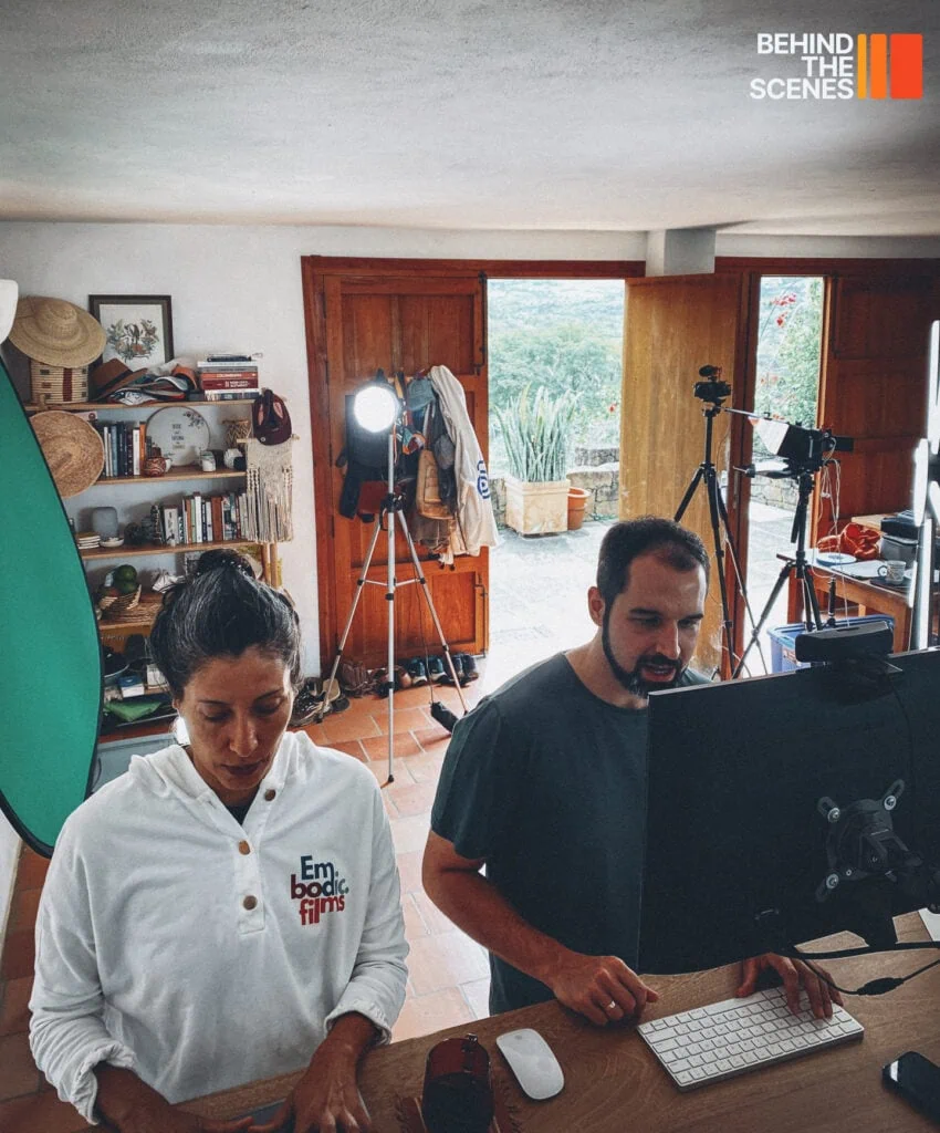 Two people work at a computer in a room set up for filming, with lights, cameras, and Embodic Films equipment visible. The "BEHIND THE SCENES" logo appears in the top right corner.
