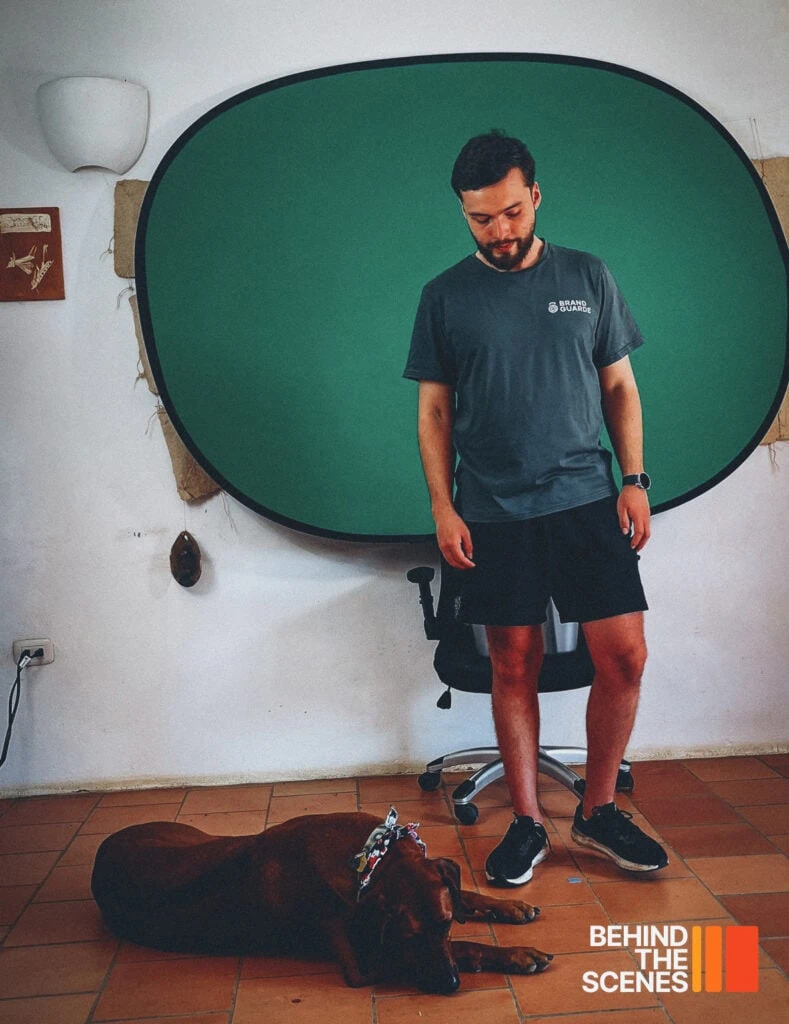 A man in a casual outfit stands in front of a green screen backdrop while a brown dog lies on the floor. "Behind the Scenes" and the Embodic Films logo are visible in the bottom right corner.