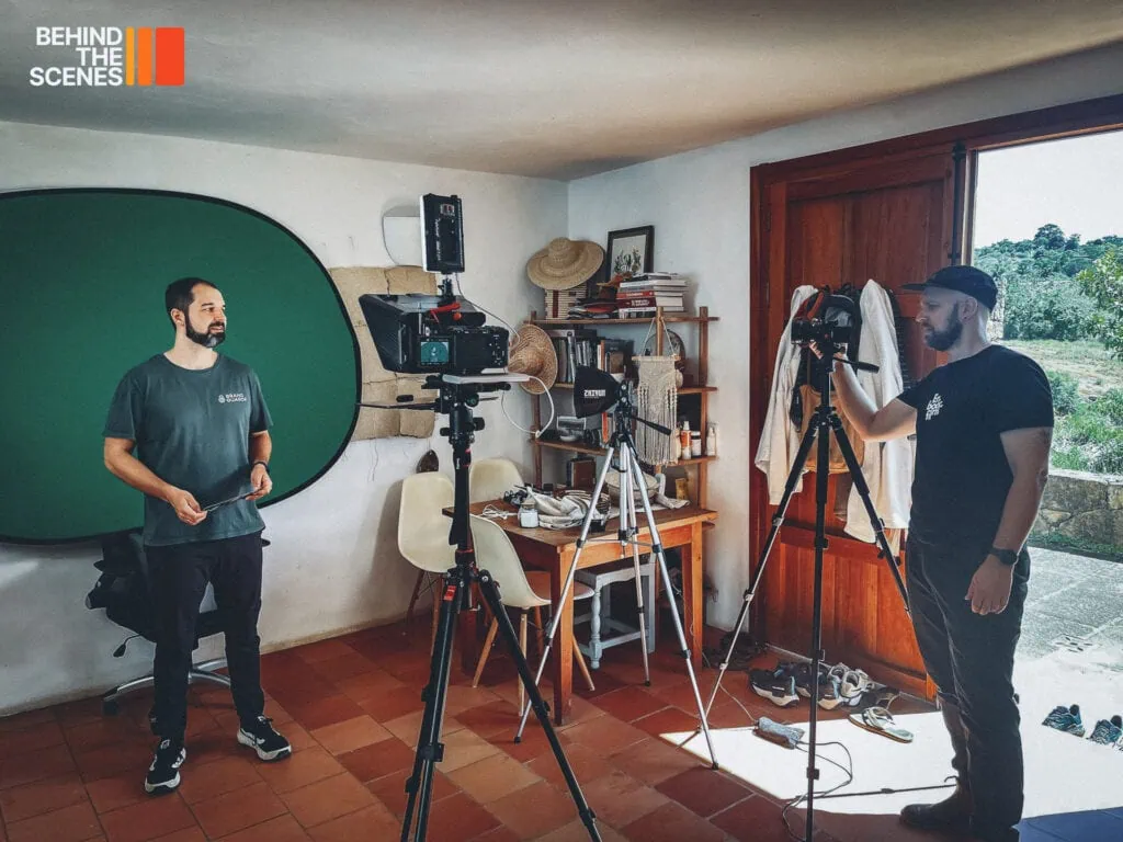 Two men from Embodic Films set up camera equipment in a small room with a green screen, desk, and scattered items, preparing for a video shoot.