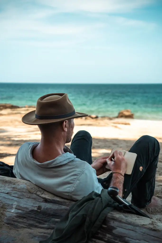 A person wearing a hat sits on a driftwood log at the beach, writing in a notebook while facing the ocean—a tranquil moment captured in true Embodic Films style.