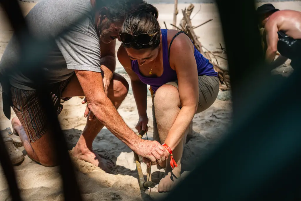 Two people kneel on sandy ground, using a stick and dry materials to start a fire, with branches nearby and a blurred figure in the background—capturing an authentic moment for Embodic Films.