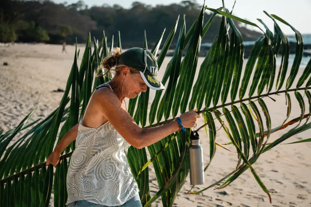A woman on a sandy beach carries large green palm fronds and holds a reusable water bottle, with trees and ocean waves in the background—a serene scene captured by Embodic Films.