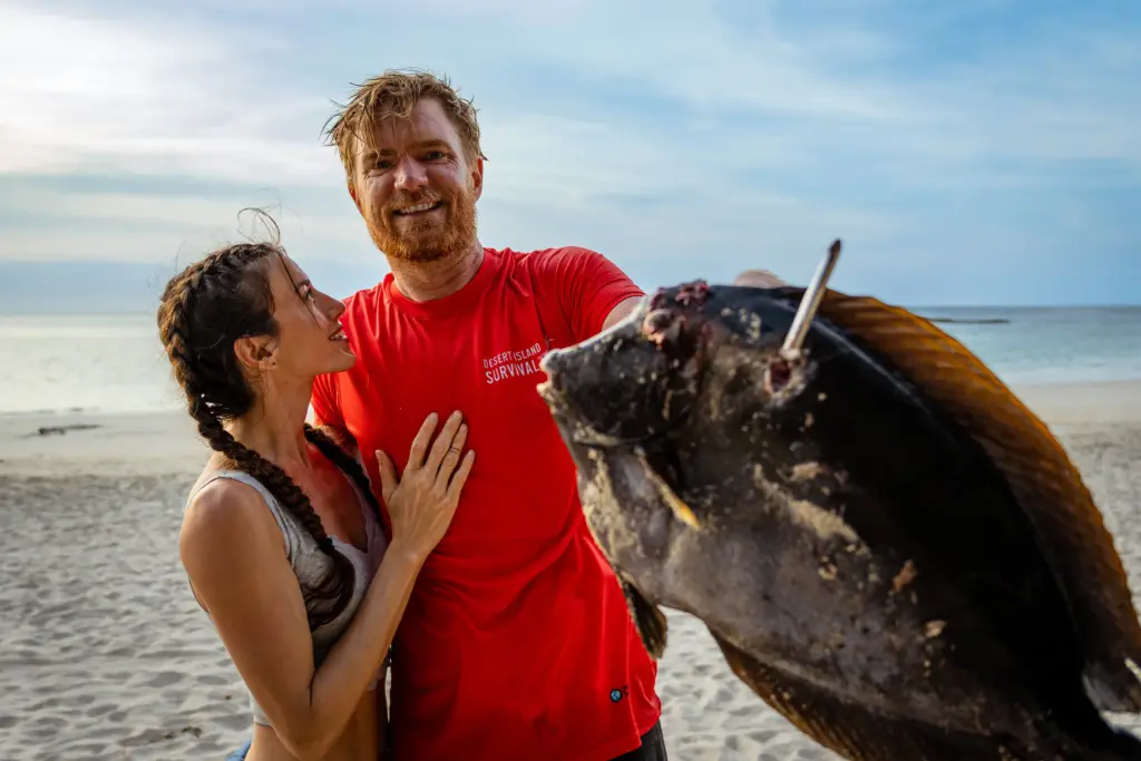 A man in a red shirt holds a large fish on a beach while a woman stands beside him, both smiling. The ocean and sky are in the background in this joyful Embodic Films moment.