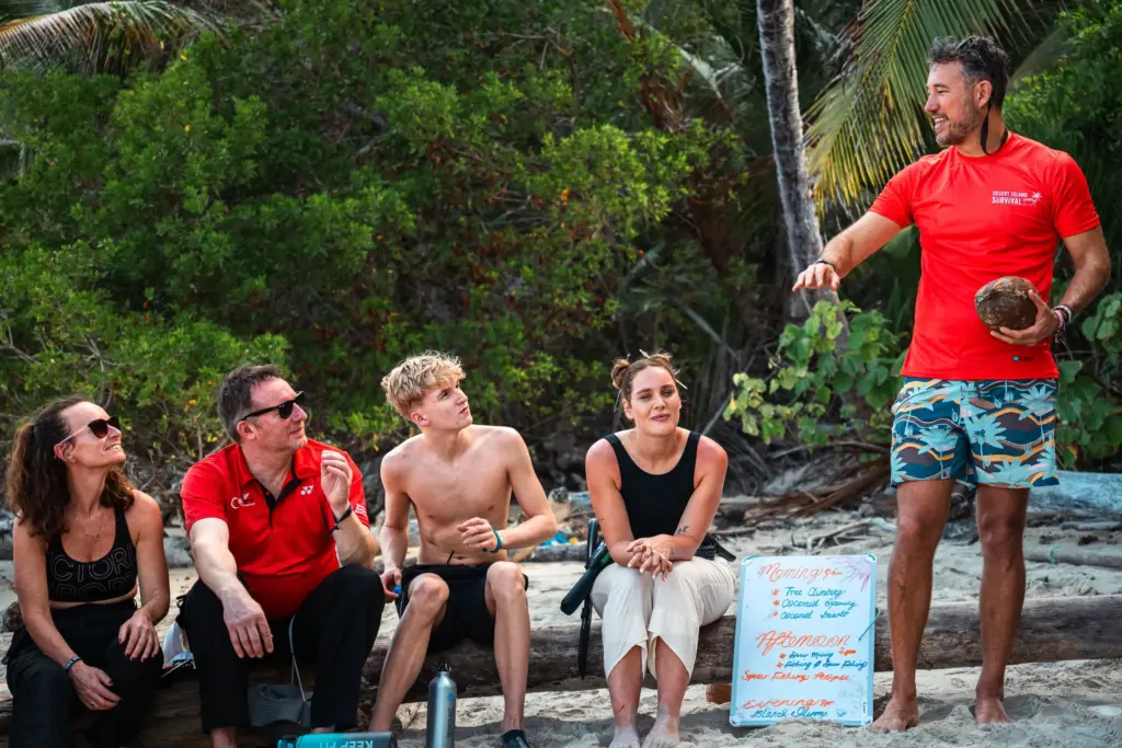 Five people sit on logs on a sandy beach; one person stands holding a coconut and appears to be speaking. A whiteboard with writing is visible in the background, capturing a behind-the-scenes moment from Embodic Films.