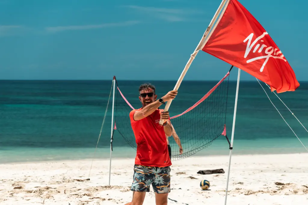 A man in sunglasses and a red shirt holds a Virgin-branded flag on a sandy beach, with a volleyball net, ocean in the background, and an Embodic Films crew capturing the lively scene.
