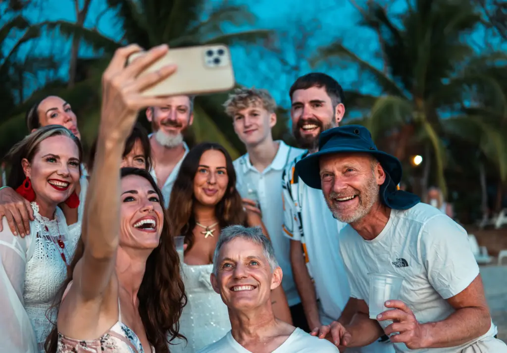 A group of people pose closely together outdoors, smiling as one person in front takes a selfie with a smartphone; Embodic Films captures the moment with palm trees swaying in the background.