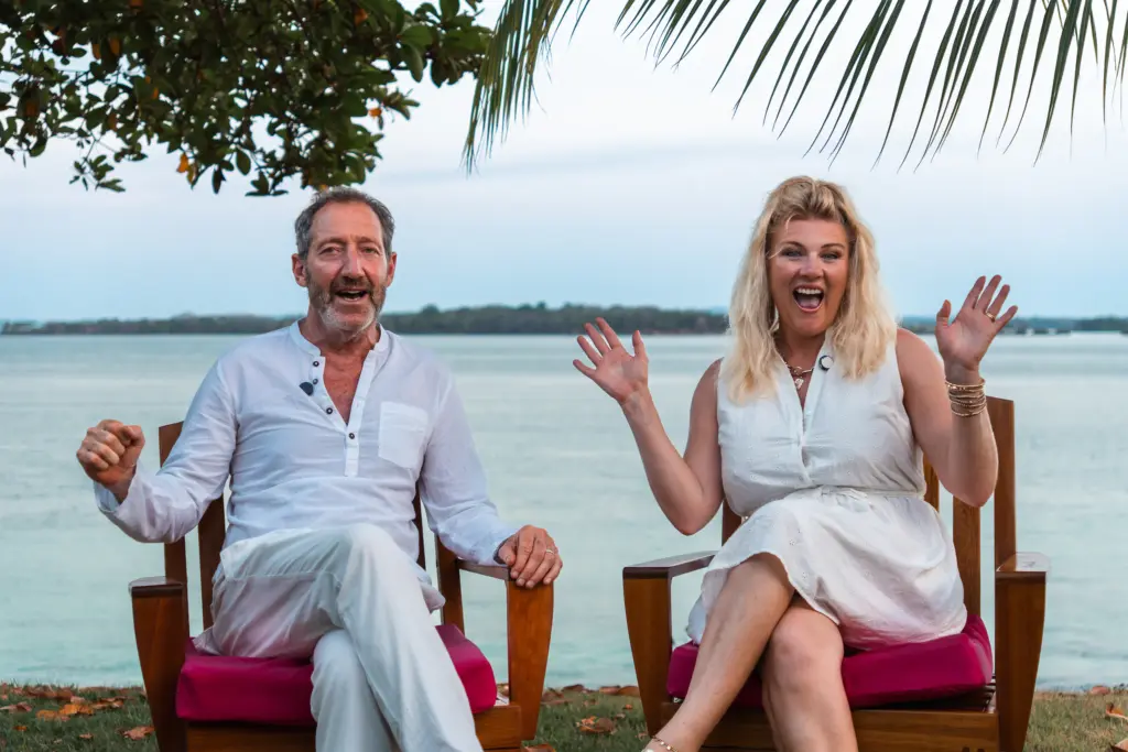 A man and a woman sit on wooden chairs by the water, both smiling and gesturing enthusiastically beneath palm leaves—a classic Embodic Films moment.