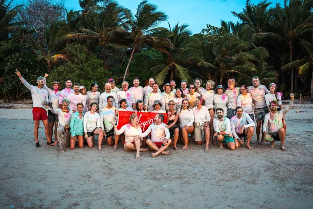 A large group of people covered in colored powder pose together on a beach with palm trees, holding a red "Virgin" banner in the center—capturing a vibrant moment with Embodic Films.