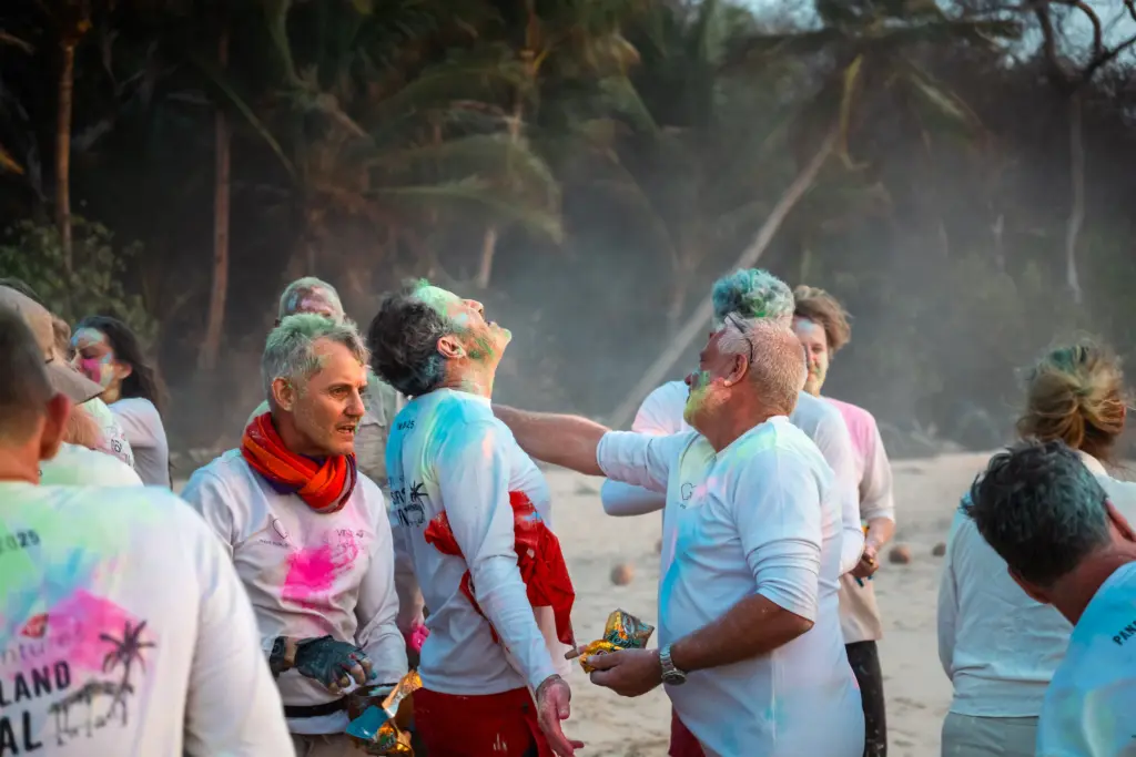 A group of people wearing white shirts stand on a beach, some with colored powder on their faces and clothes, engaging with each other in a festive atmosphere—captured beautifully by Embodic Films.