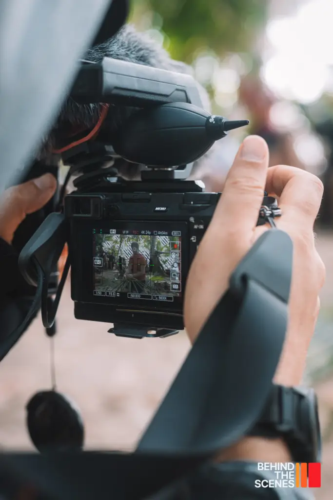 Close-up of a person holding a professional video camera, with a screen displaying an outdoor scene; "BEHIND THE SCENES" text appears in the corner, capturing the filmmaking process with Embodic Films.