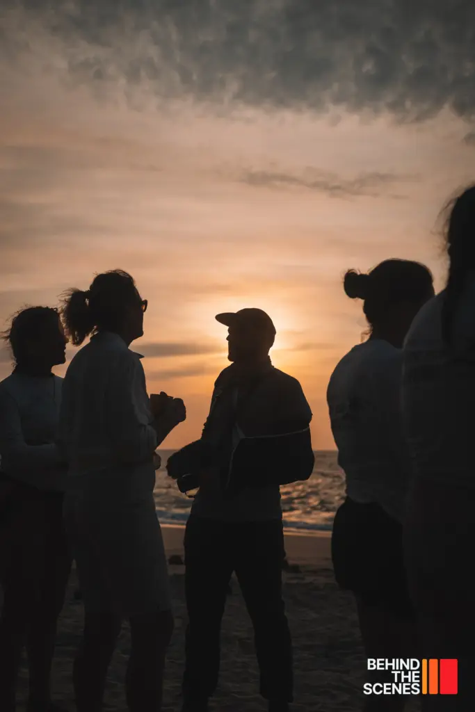 A group of people stand and talk on a beach at sunset, appearing as silhouettes against the sky and ocean, with "Behind the Scenes" and Embodic Films text in the bottom right corner.