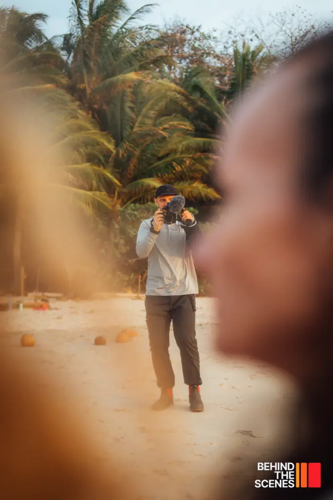 A person stands on a sandy beach holding a camera, with palm trees in the background and a blurred figure in the foreground. The text "Behind the Scenes" and "Embodic Films" appears in the bottom right corner.