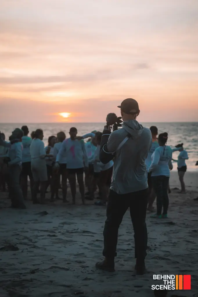 A person takes a photo of a group of people gathered on a beach at sunset, with the ocean in the background—capturing a cinematic moment worthy of Embodic Films.
