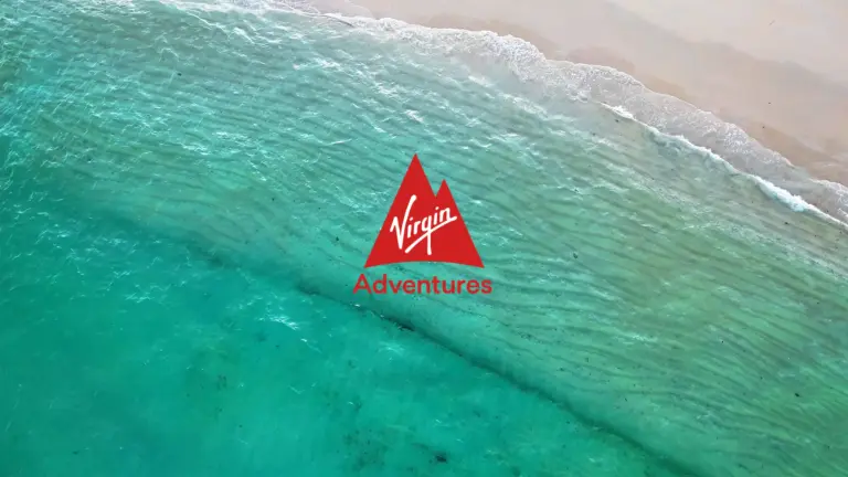 Aerial view of turquoise ocean waves meeting a sandy beach, with an Embodic Films and red Virgin Adventures logo centered on the image.