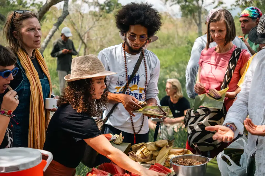 A group of people gather outdoors around a table covered with food, including tamales. Some are serving themselves, while others chat and reach for items, enjoying a casual, communal meal in a natural setting.