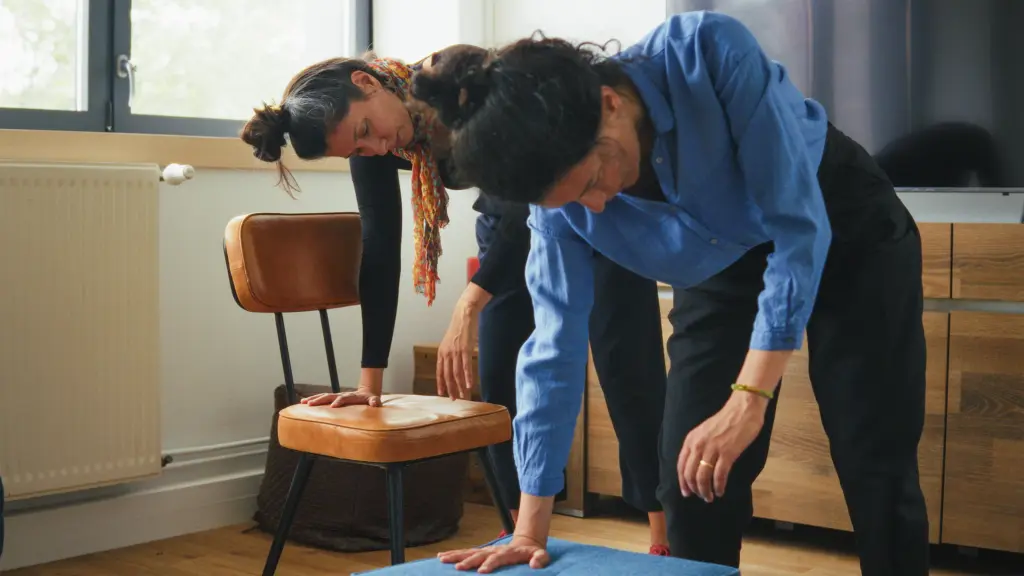 Two women perform a stretching exercise indoors, each leaning forward with one hand on a supportive surface and the other arm extended downward.