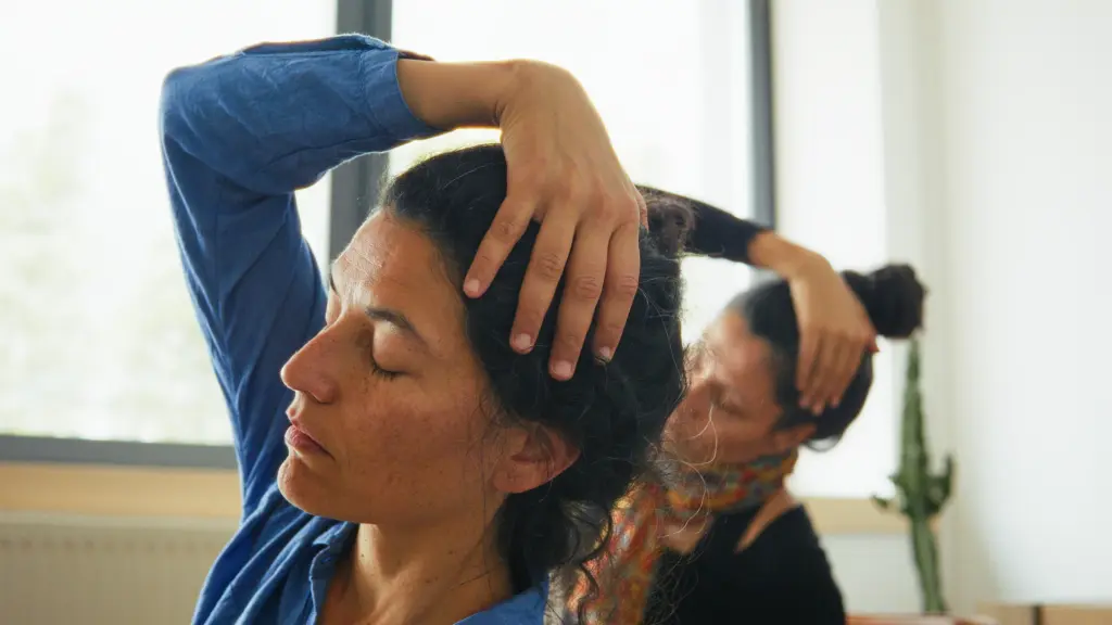 Two women sit indoors, eyes closed, each using one hand to gently stretch their necks to the side.