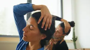 Two women sit indoors, eyes closed, each using one hand to gently stretch their necks to the side.