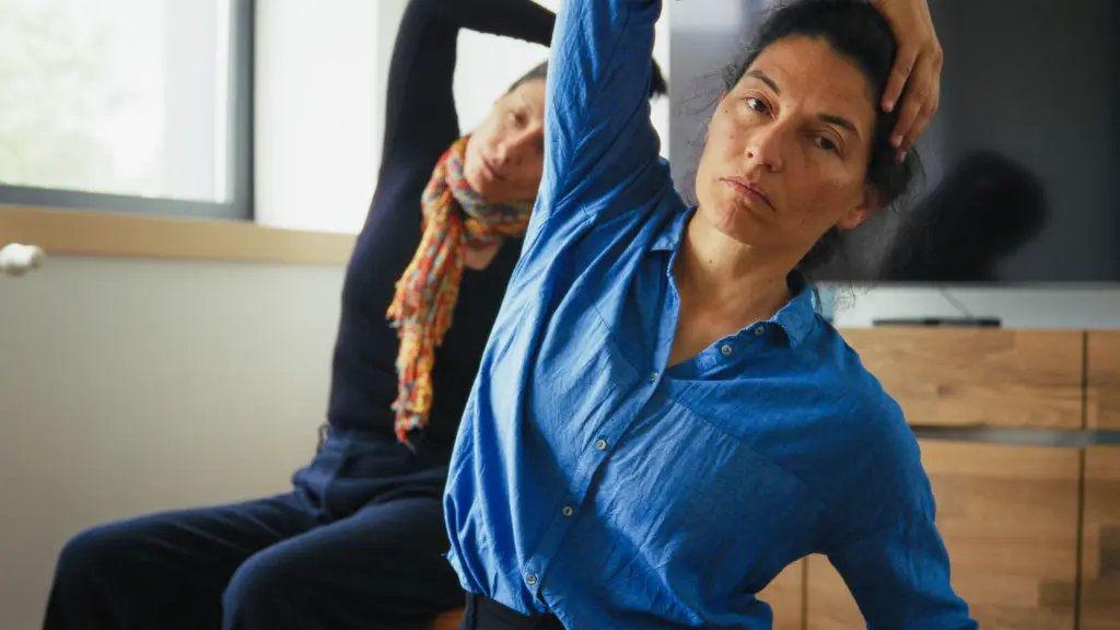 Two women seated indoors perform a side stretch, each with one arm raised over their head. One wears a blue shirt, the other a patterned scarf.