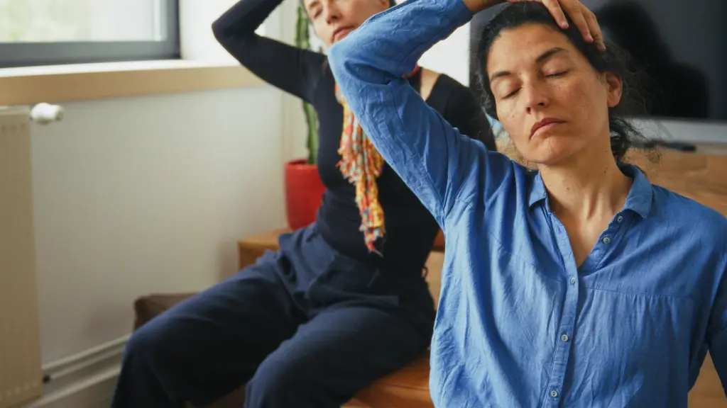 Two people seated indoors with eyes closed, gently stretching their necks by tilting their heads to one side and using a hand to assist the stretch.
