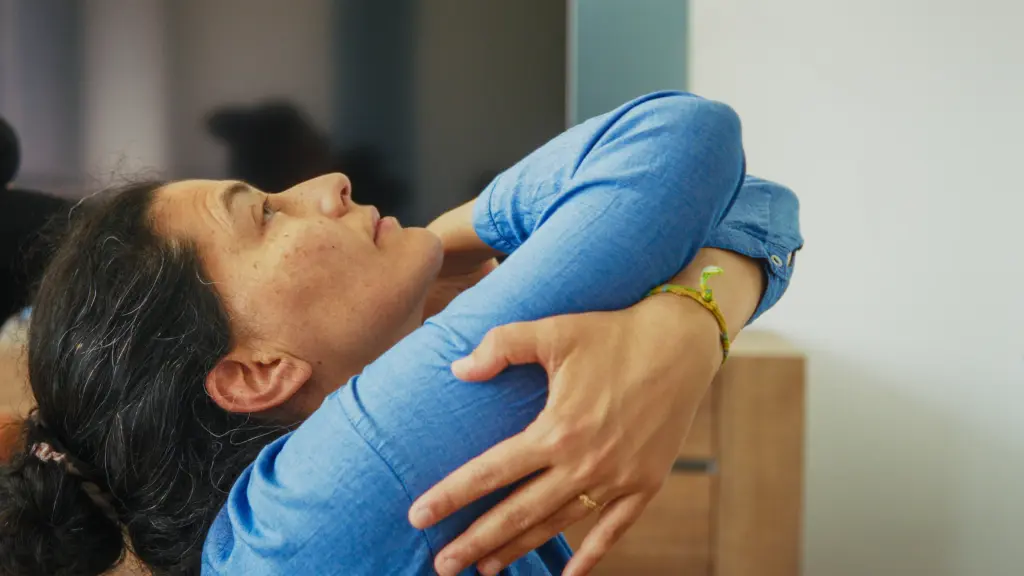 Woman in a blue shirt sits sideways, crossing her arms and stretching her back while tilting her head back, with eyes closed.