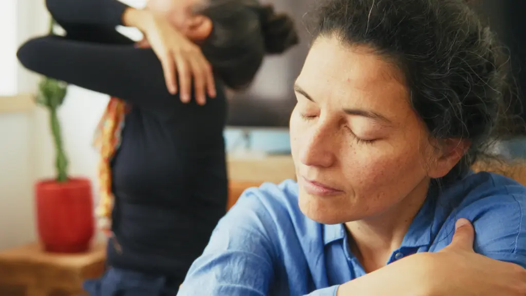 Two women with closed eyes sit indoors, each resting a hand on their own shoulder. One is in focus wearing a blue shirt; the other is blurred in the background near a red plant pot.