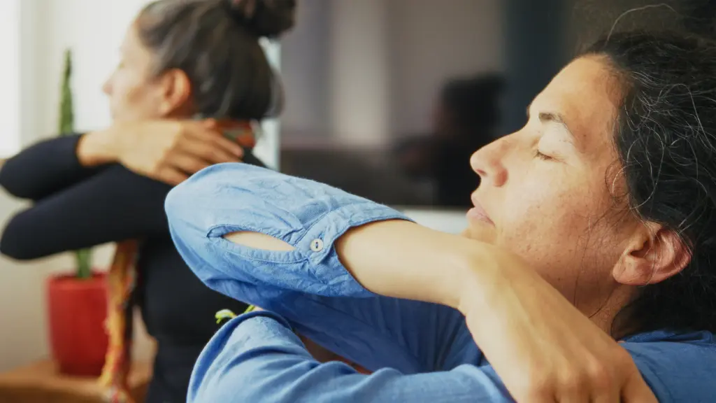 Two women with closed eyes practice self-hugging or self-soothing gestures indoors, with a plant visible in the background.