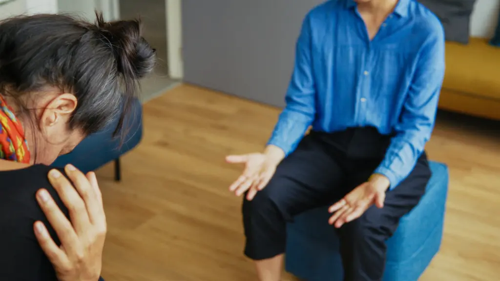 Two people sit indoors; one person leans forward with their head down, while the other gestures with open hands, suggesting a conversation or counseling session.