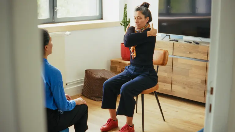 A woman sitting on a chair crosses her arms over her chest, while another person sits nearby observing in a living room.