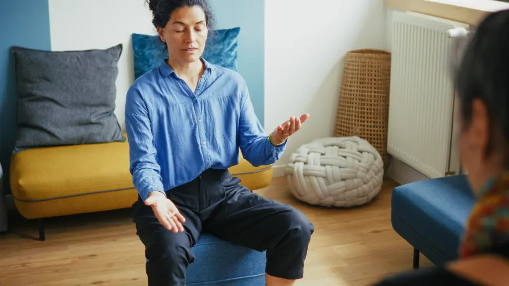 A person sits on a blue chair with eyes closed and arms open, appearing to meditate or practice mindfulness in a cozy, modern room.