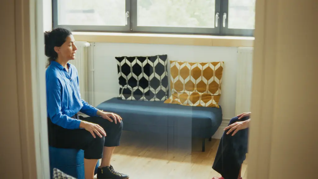Two people sit facing each other in a brightly lit room with geometric-patterned cushions on a bench and large windows in the background.