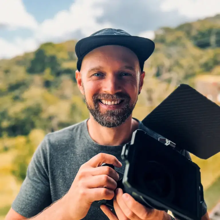 Smiling man with beard holding camera and light in outdoor setting.