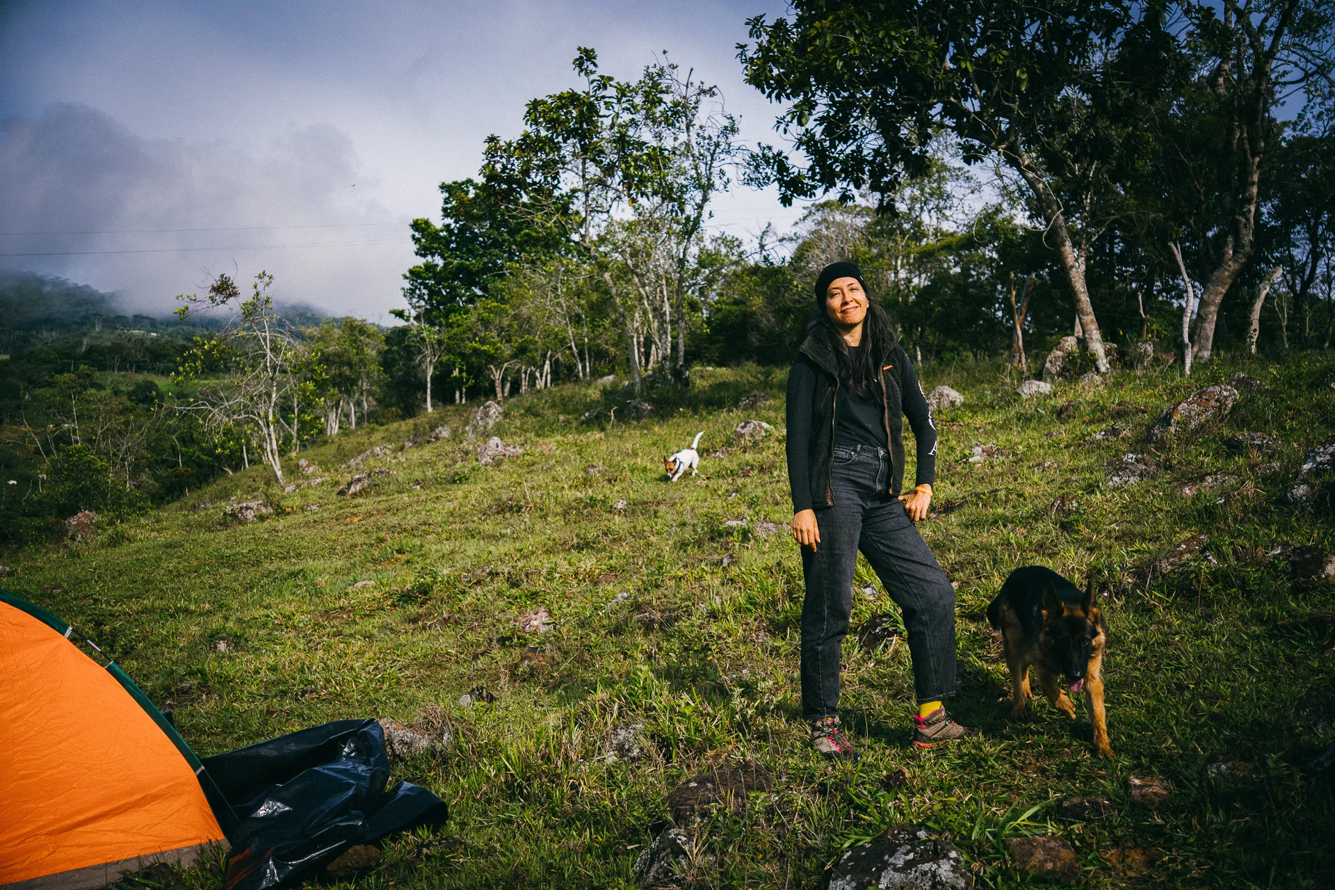 Woman camping in El Páramo with dogs, wearing a black jacket and jeans. Tent visible. Cloudy sky, green landscape.