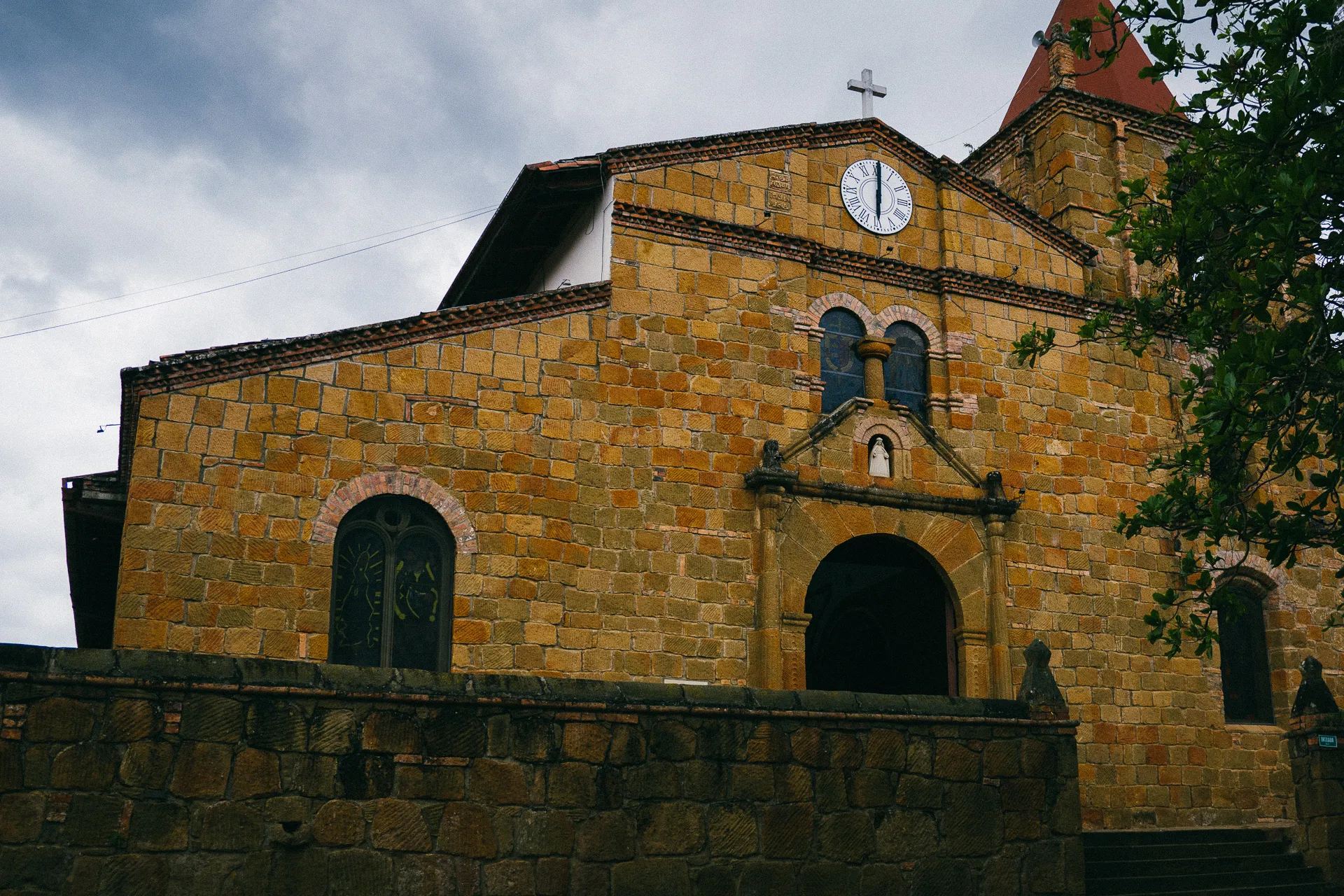 Facade of a yellow stone church with a clock and arched doorway under a cloudy sky.